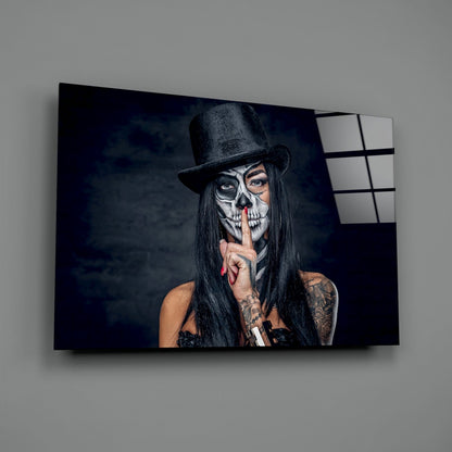 Glass wall art displaying a person in skull makeup and black top hat with finger to lips against dark background, mounted on gray wall.