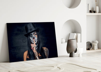 Glass wall art leaning against white wall displaying person with skull face paint, black hat, and finger to lips in minimalist interior setting.