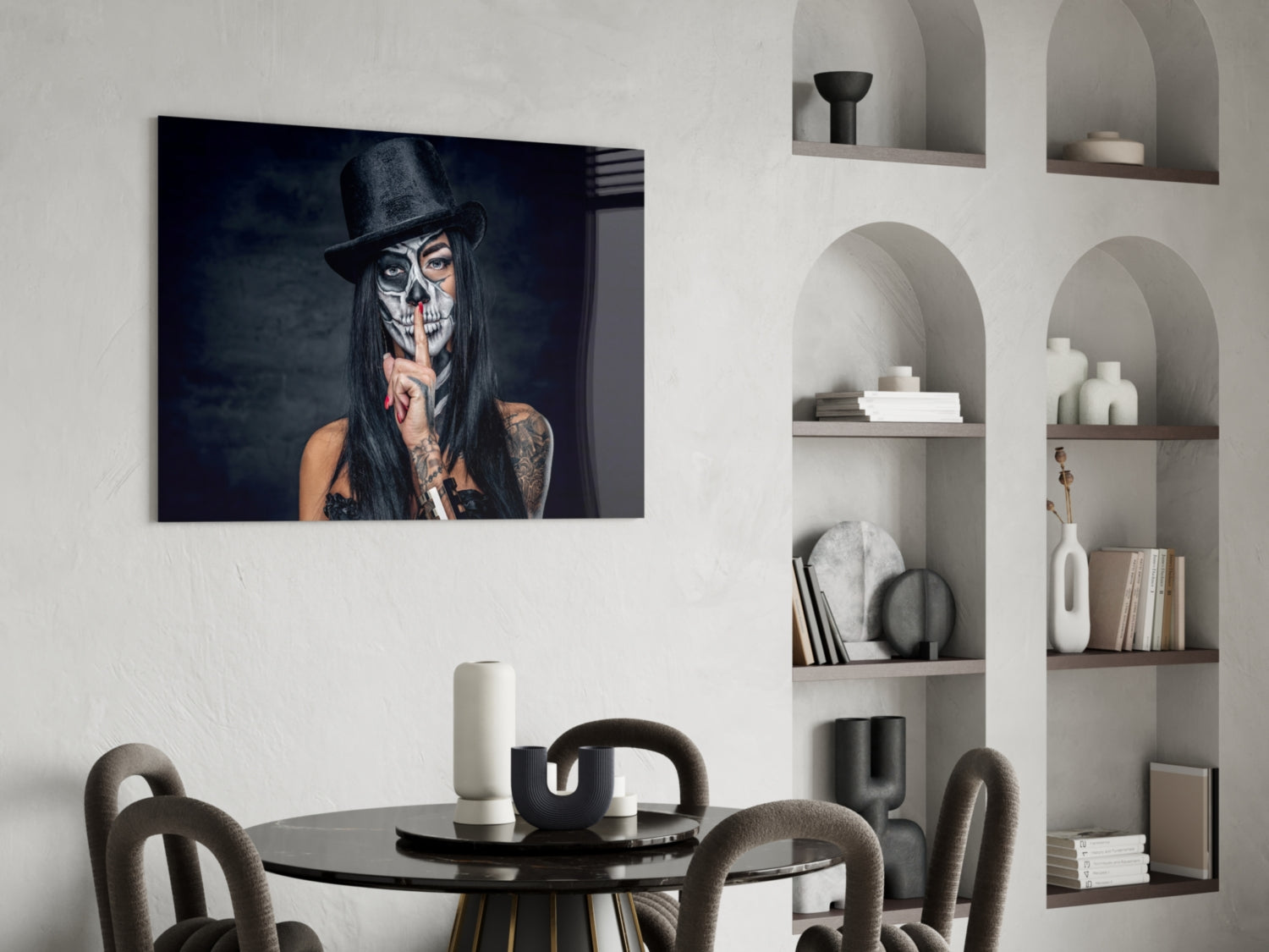 Skull-faced figure wearing black top hat with finger to lips, displayed as framed glass wall art above round black dining table.