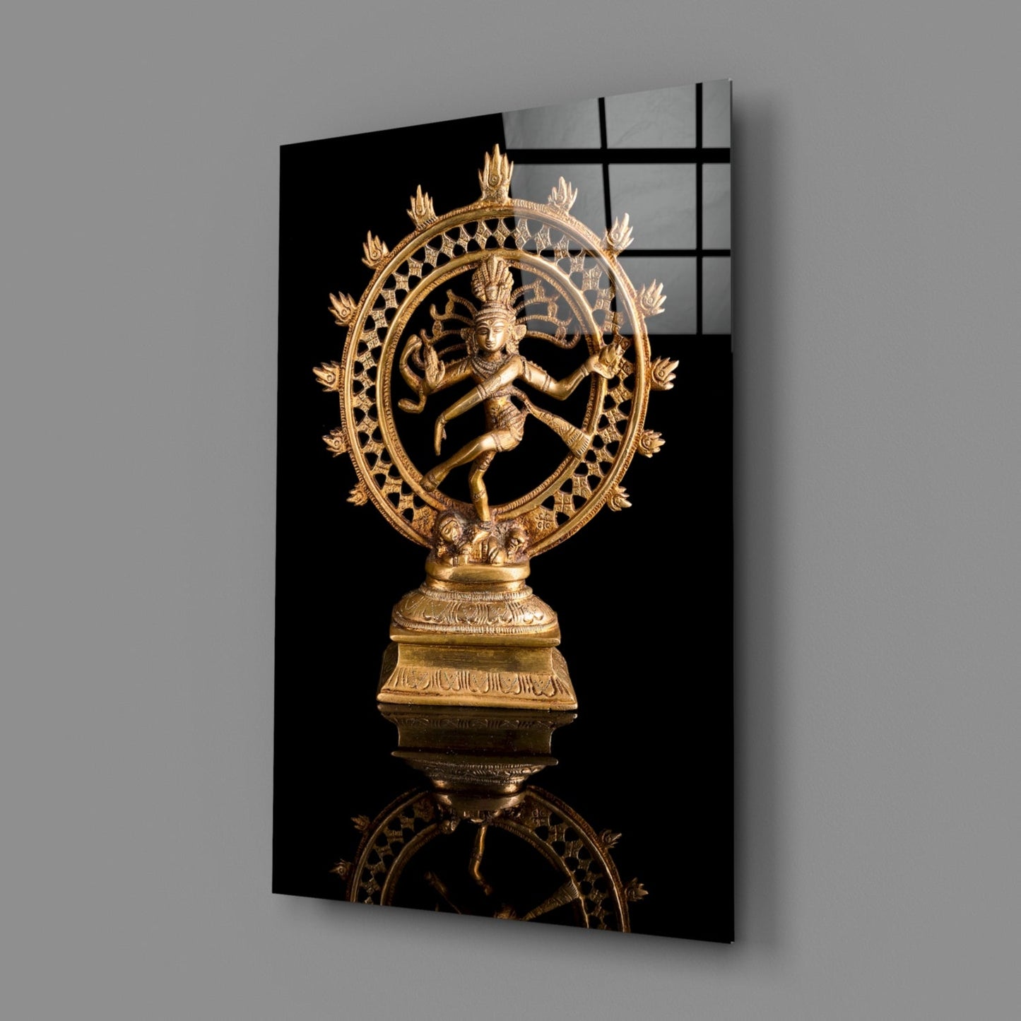 Golden Shiva Nataraja statue mounted on black glass panel, displayed at angle against gray wall showing reflection.