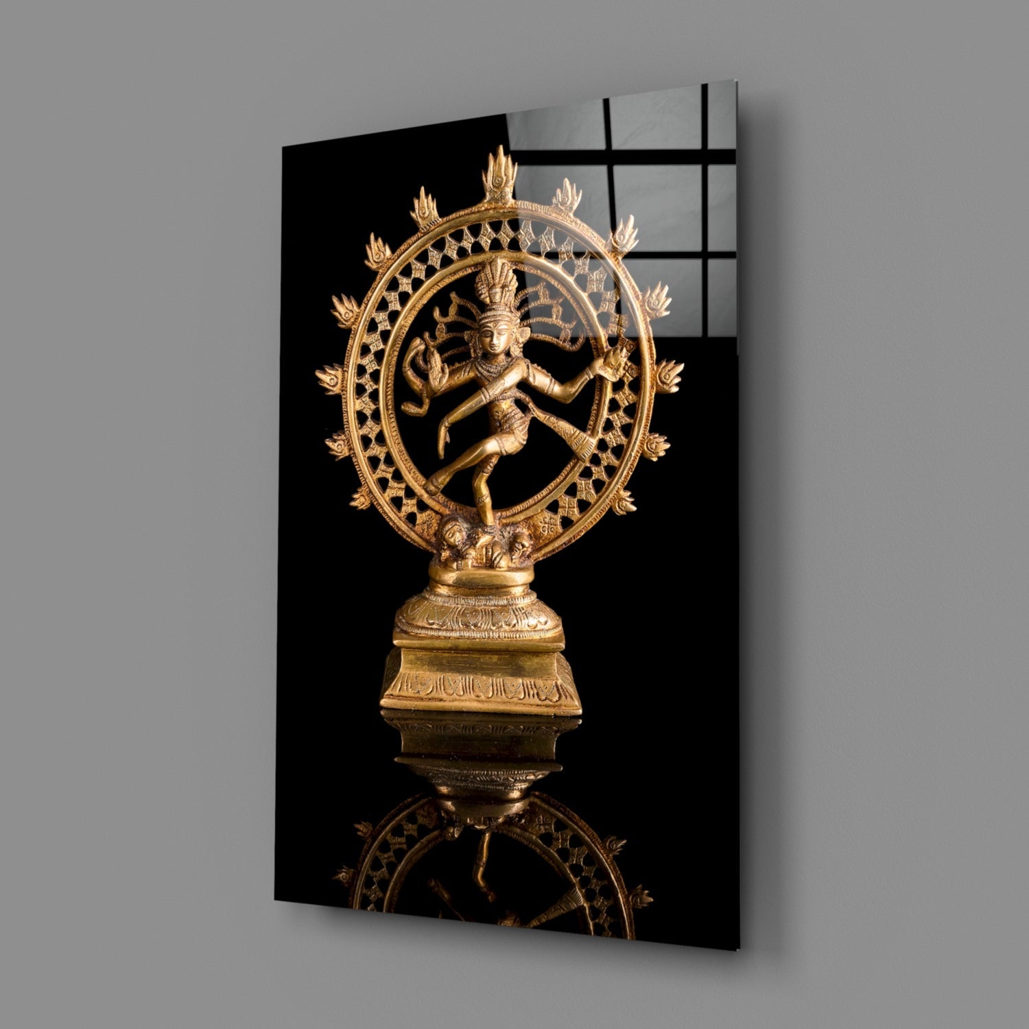 Golden Shiva Nataraja statue mounted on black glass panel, displayed at angle against gray wall showing reflection.