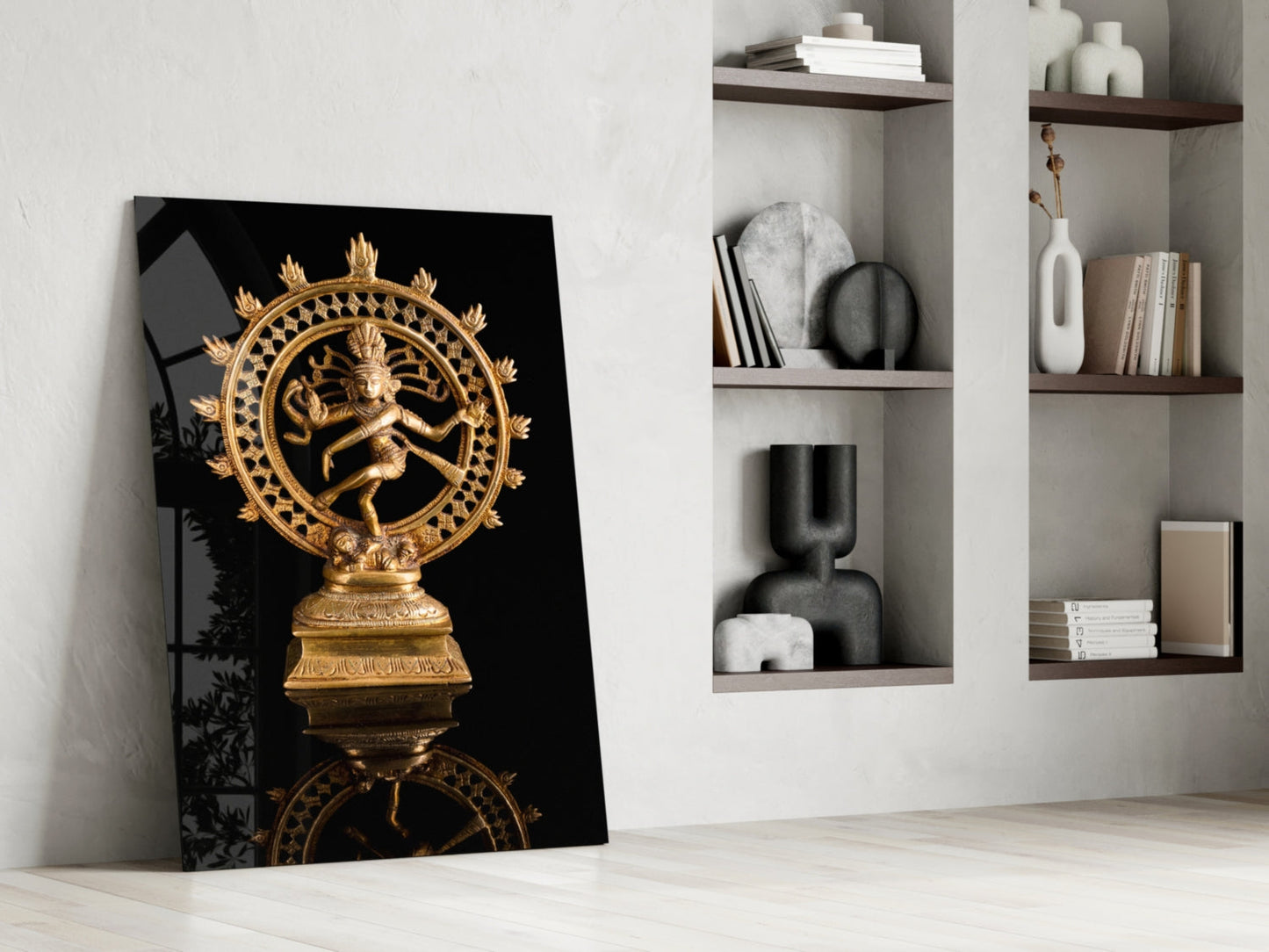 Golden Shiva Nataraja statue on black glass panel leaning against white wall beside wooden shelving.
