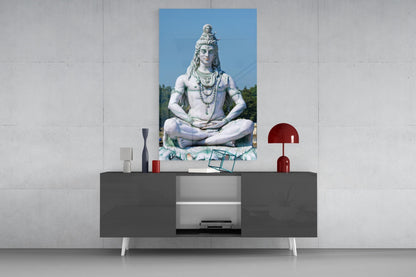 Glass wall art depicting a white stone Shiva statue in meditation pose displayed above a dark console table with decorative accessories.