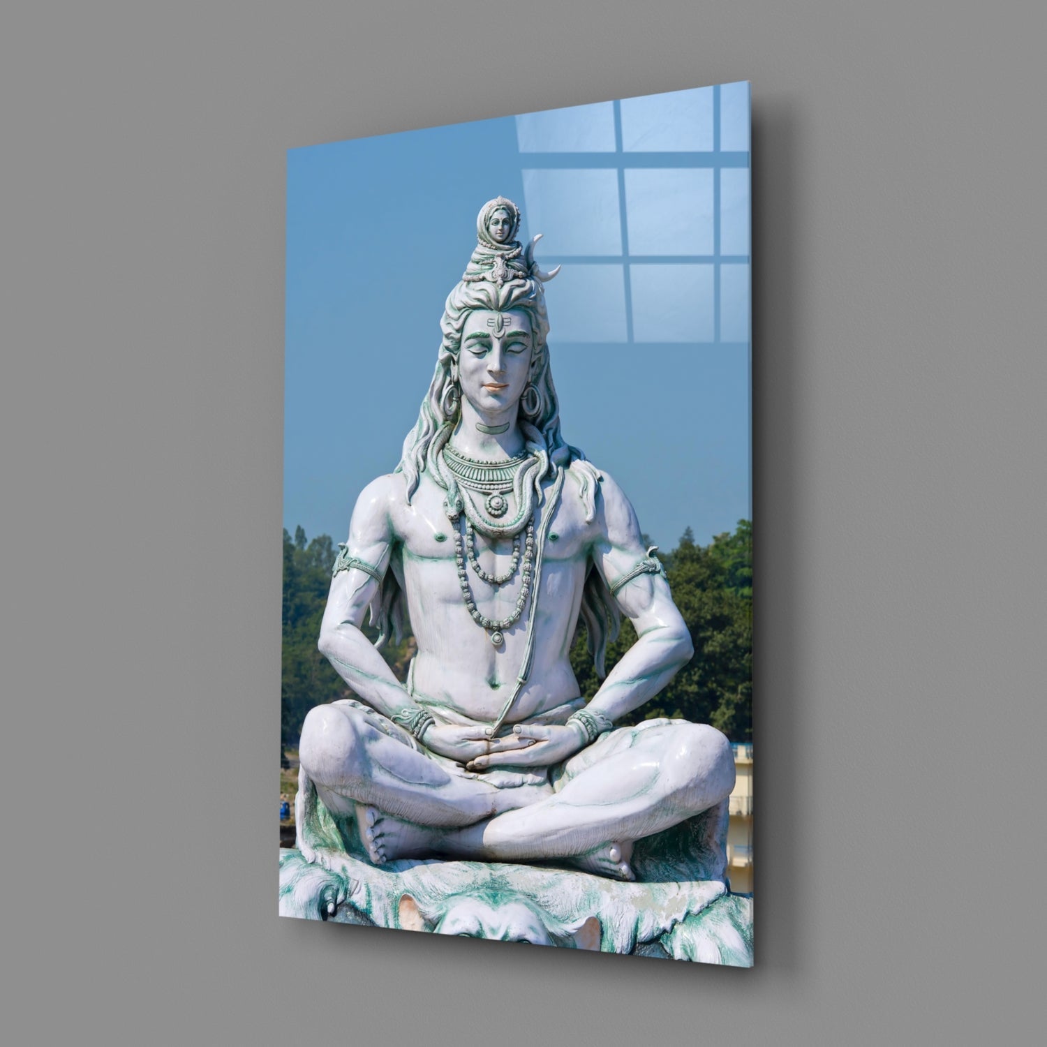 Large white marble statue of seated Shiva in meditative pose with ornamental jewelry, displayed on glass wall art panel against blue sky background.