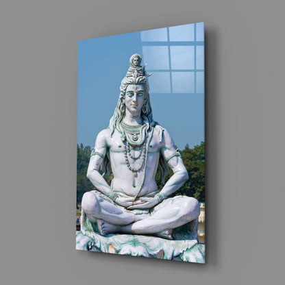Large white marble statue of seated Shiva in meditative pose with ornamental jewelry, displayed on glass wall art panel against blue sky background.
