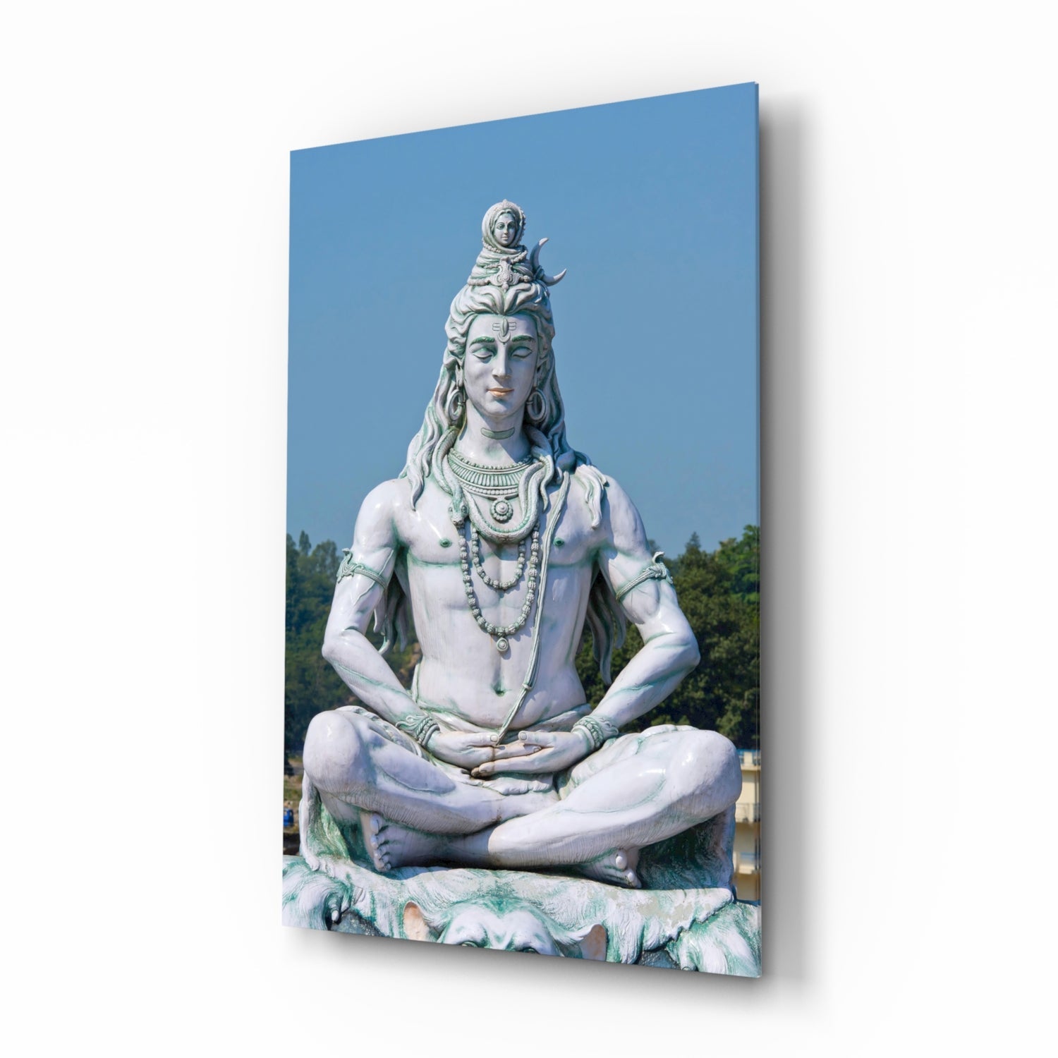 Large white stone statue of meditating Shiva with ornate headdress and jewelry, seated in lotus position against blue sky with forested background.