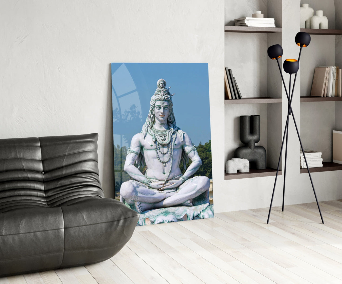 Glass wall art depicting Lord Shiva meditating in white and blue tones, displayed in a modern living room beside black furniture and shelving.