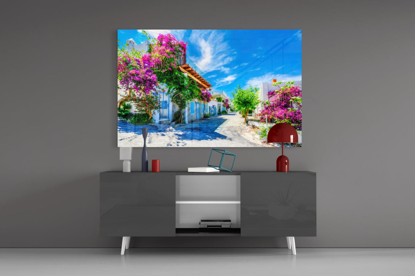 Glass wall art displaying colorful Mediterranean street scene with pink bougainvillea flowers mounted above gray television console.