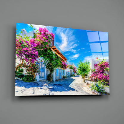Glass wall art mounted on gray wall displaying Mediterranean street scene with white buildings, blue doors, and vibrant magenta bougainvillea flowers.