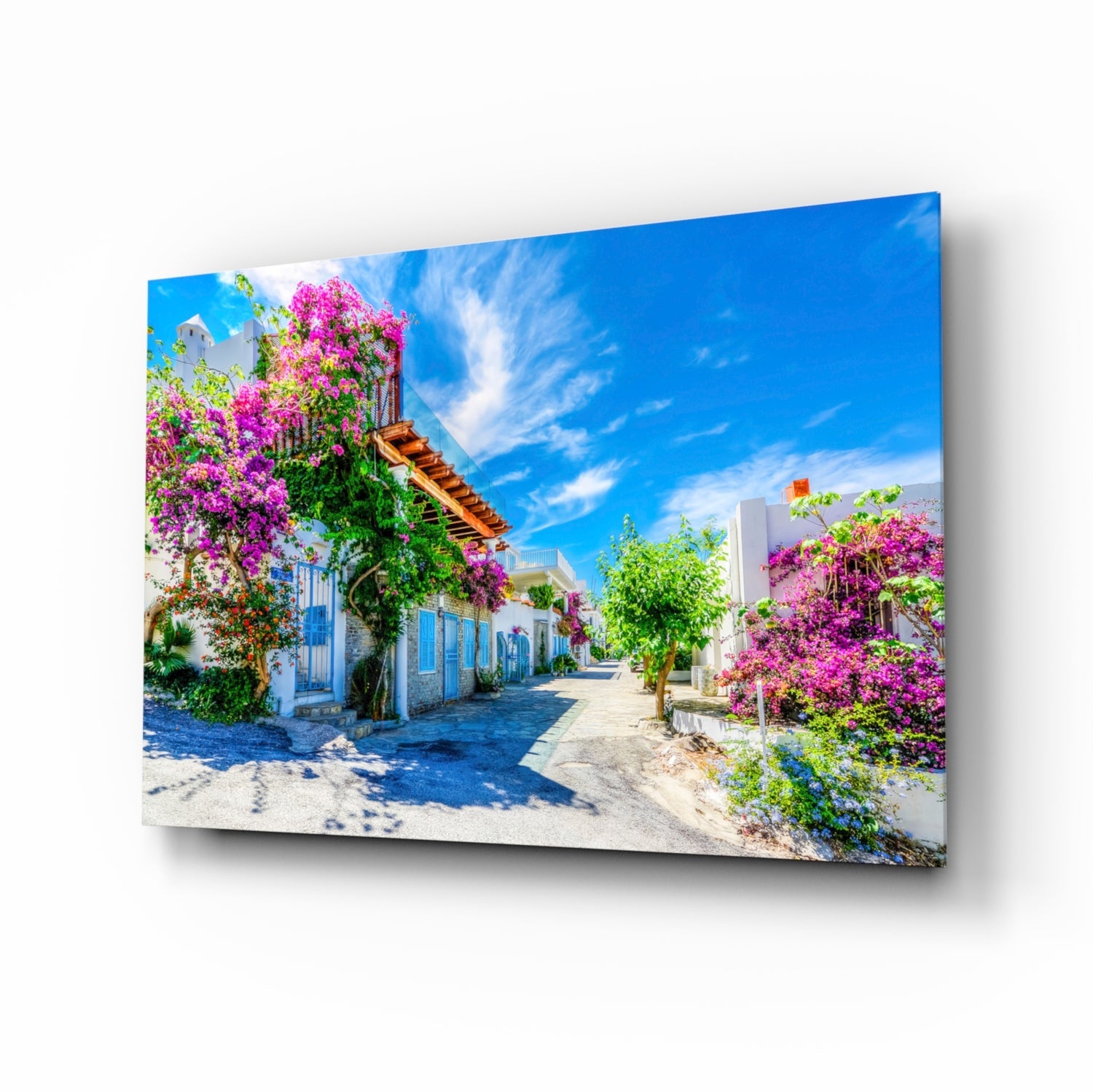 Glass wall art displaying a Mediterranean village street with white-walled buildings, vibrant magenta bougainvillea flowers, and blue sky.