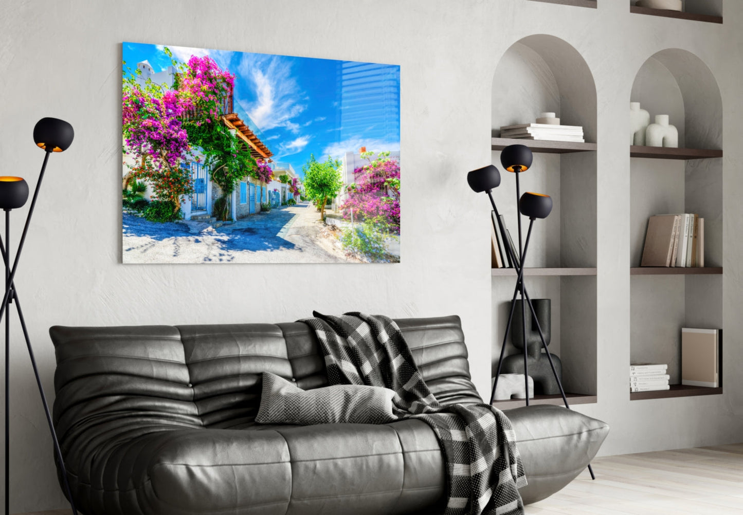Glass wall art print of a Mediterranean street scene with pink flowering bougainvillea and blue buildings, displayed above a black leather sofa.