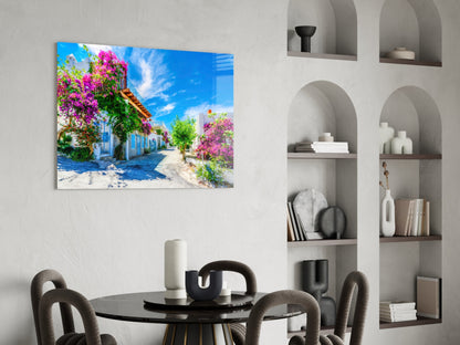 Colorful glass wall art depicting a Mediterranean street with magenta bougainvillea flowers and blue sky, displayed above a black dining table.