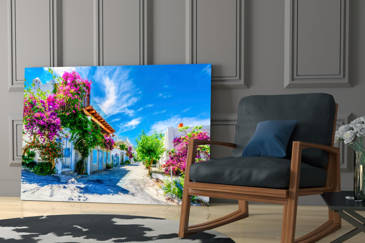 Aegean Beauties glass wall art displayed in modern interior with wooden chair, showing vibrant Mediterranean street scene with pink bougainvillea flowers and blue architecture.