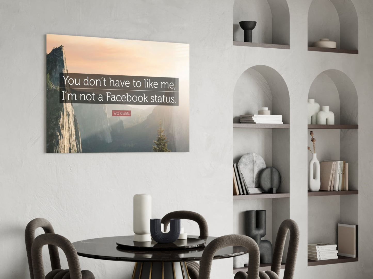 Glass wall art with mountain landscape photograph and white text quote displayed above round black dining table with gray chairs.