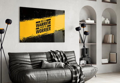 Black leather sofa against white wall with yellow and black "Be a Warrior Not a Worrier" glass art hanging above it, flanked by arched shelving and black floor lamps.