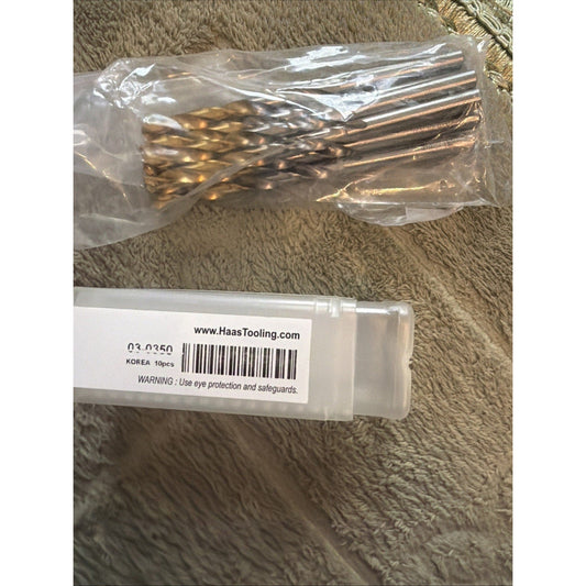 Packaging and contents of Haas Tooling drill bit set showing labeled box and clear plastic bag containing gold-colored metal bits on tan fabric background.