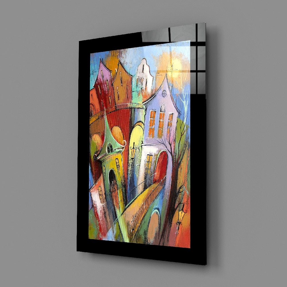 Angled view of colorful glass wall art depicting vibrant whimsical buildings with yellow, red, pink, and green hues in a black frame.