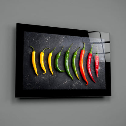 Glass wall art displaying nine chili peppers in gradient color progression from yellow to red on dark textured background.