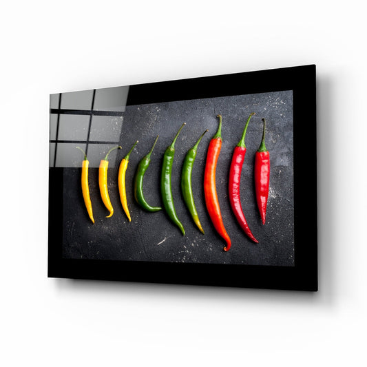 Black-framed glass wall art displaying nine chili peppers arranged in gradient from yellow to red on dark textured background, angled perspective view.