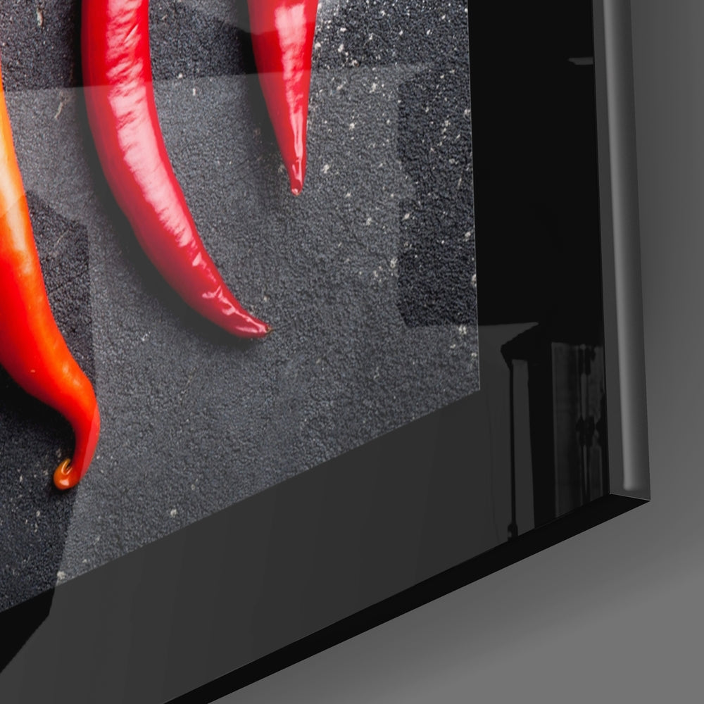 Close-up angled view of glass wall art displaying red and orange peppers on dark textured background, showing edge detail and mounting frame.
