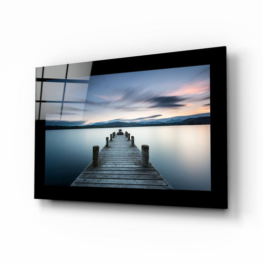 Black-framed glass wall art displaying a wooden jetty extending into calm blue water at sunset, mounted at an angle showing reflection.
