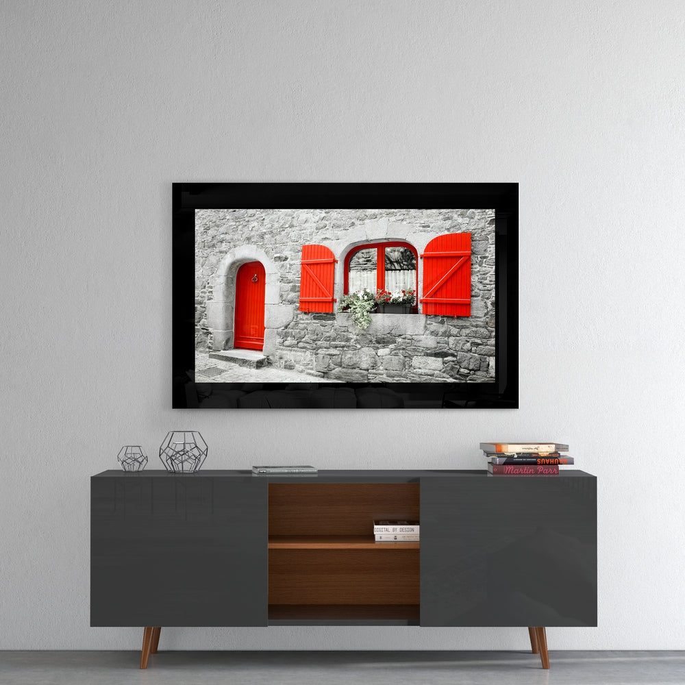 Framed glass wall art displaying a stone cottage facade with bright red door, shutters, and window frame against gray masonry.