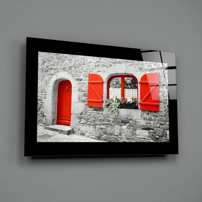 Black-framed glass wall art displaying a stone cottage facade with bright red door, shutters, and window frames on gray wall.