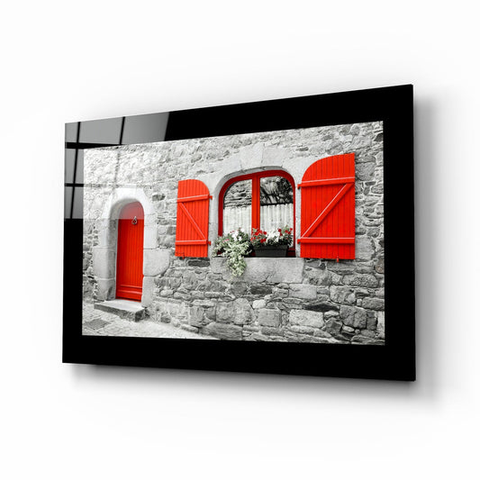 Framed wall art displayed at an angle showing a stone building facade with red door and shutters against white stonework.