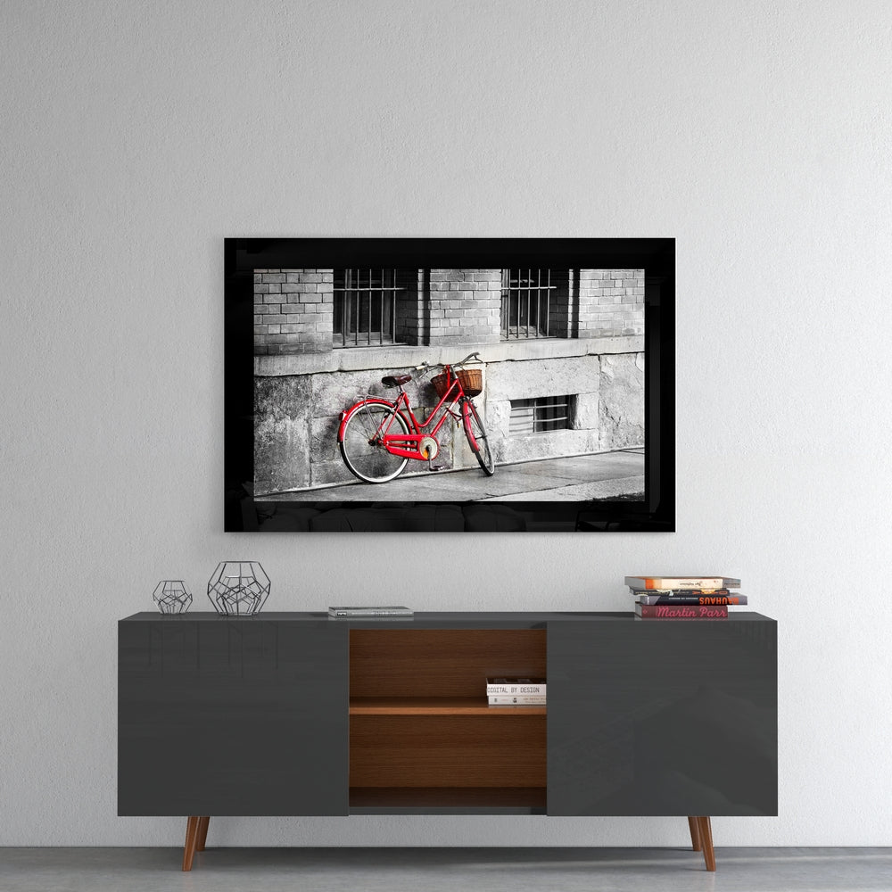 Red vintage bicycle with basket leaned against weathered gray building wall, framed in black glass.
