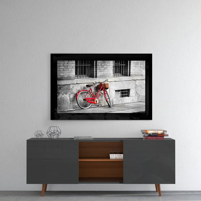 Red vintage bicycle with basket leaned against weathered gray building wall, framed in black glass.
