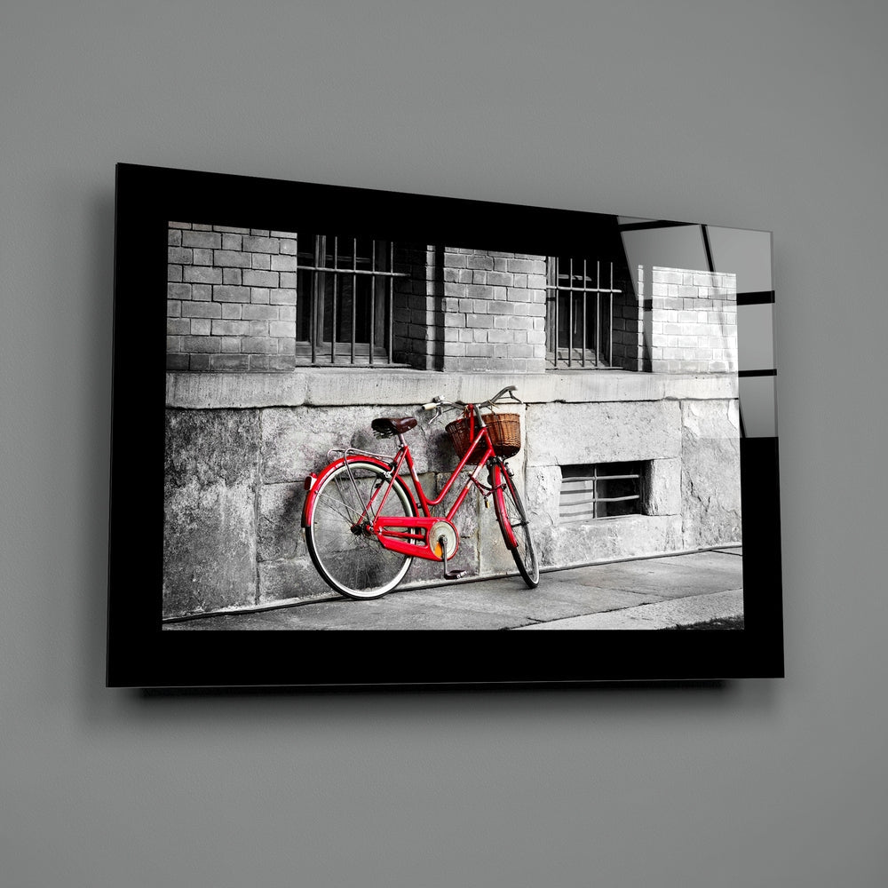 Red bicycle with basket leaned against weathered gray building wall, displayed in black-framed glass wall art.
