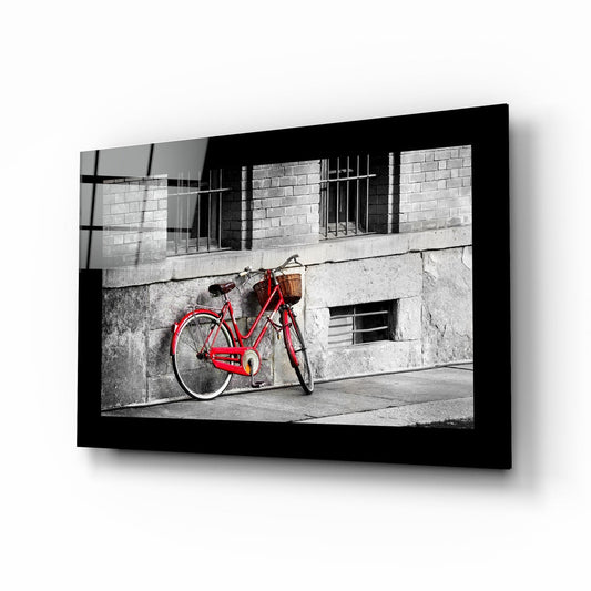 Red vintage bicycle with wicker basket leaned against weathered gray building wall in black-framed glass art display.