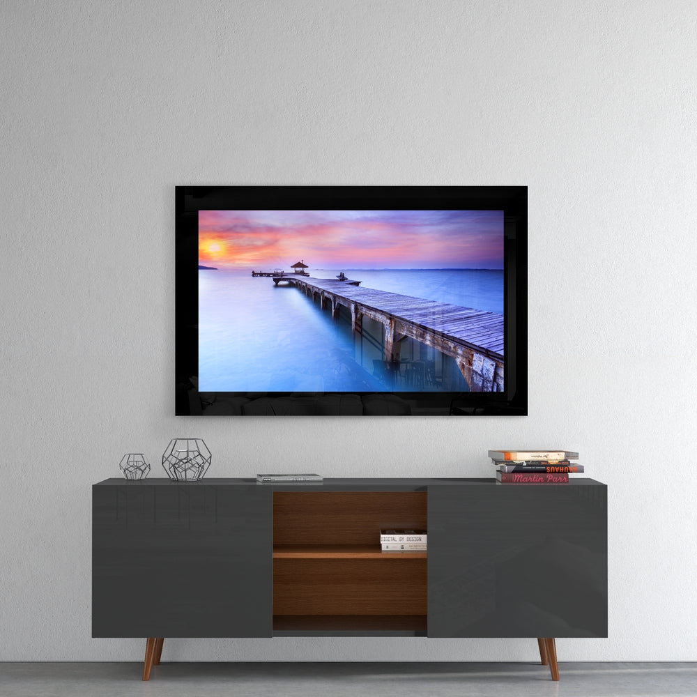 Framed glass wall art displaying a sunset pier scene with pink and blue sky, mounted above a mid-century modern TV console.