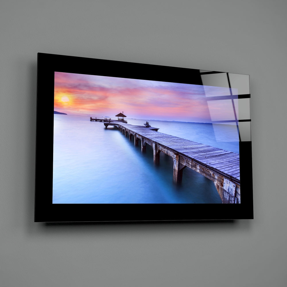 Modern glass wall art mounted on gray wall displaying a long wooden pier extending into calm blue water at sunset with golden and pink sky.