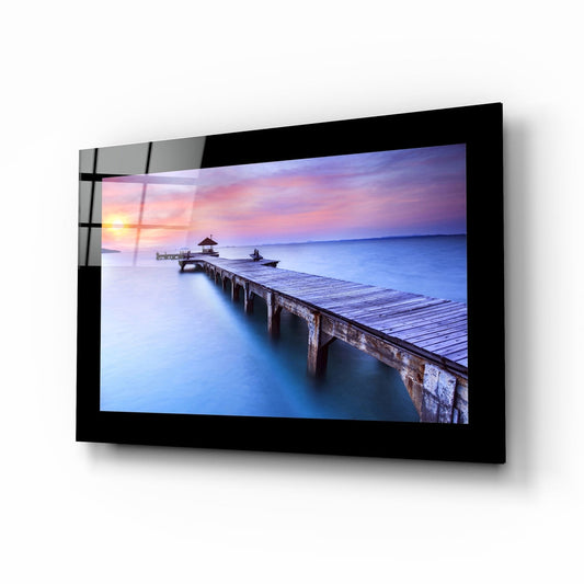 Black-framed glass wall art displaying a wooden pier extending into blue water at sunset with pink and purple sky, mounted at an angle.
