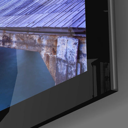Glass wall art display mounted at an angle showing weathered wooden pier pilings and blue water.