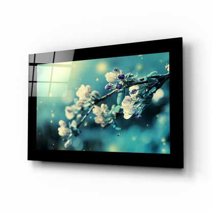 Black-framed glass wall art displaying delicate white flowers on a branch against a turquoise bokeh background, angled perspective view.