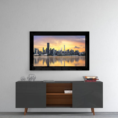 Black framed glass wall art displaying sunset cityscape with golden sky and water reflections, mounted above mid-century modern console table.