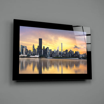 Black-framed glass wall art displaying a sunset cityscape photograph with golden sky reflected in water, mounted on gray wall.