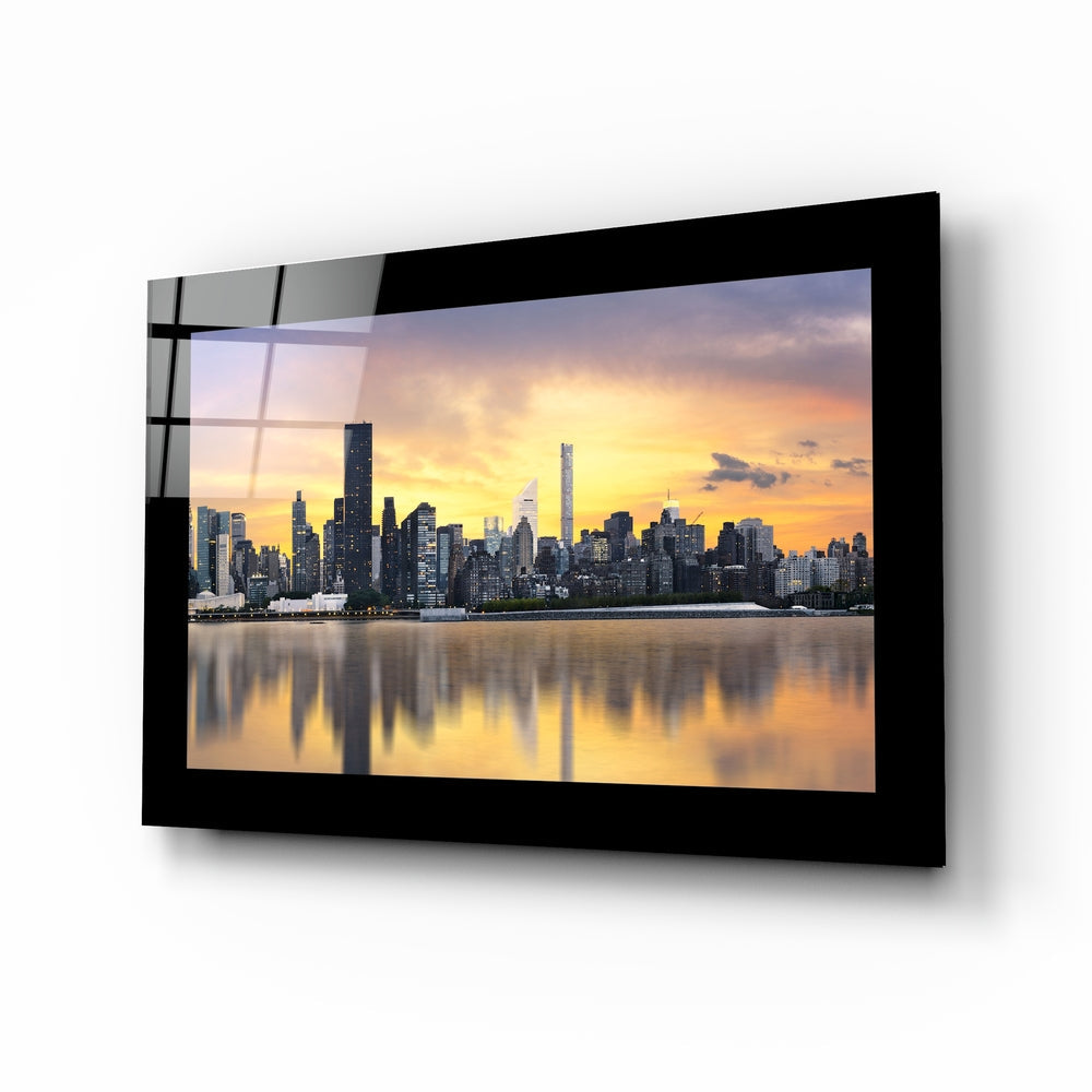 Black-framed glass wall art displaying Manhattan skyline at golden sunset reflected in still water, mounted at an angle.