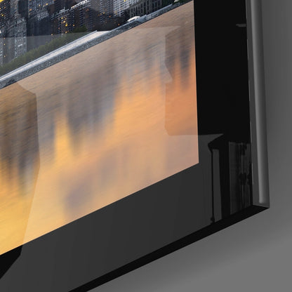 Black-framed glass wall art displaying a cityscape photograph with warm golden and orange sunset tones, mounted at an angled perspective.