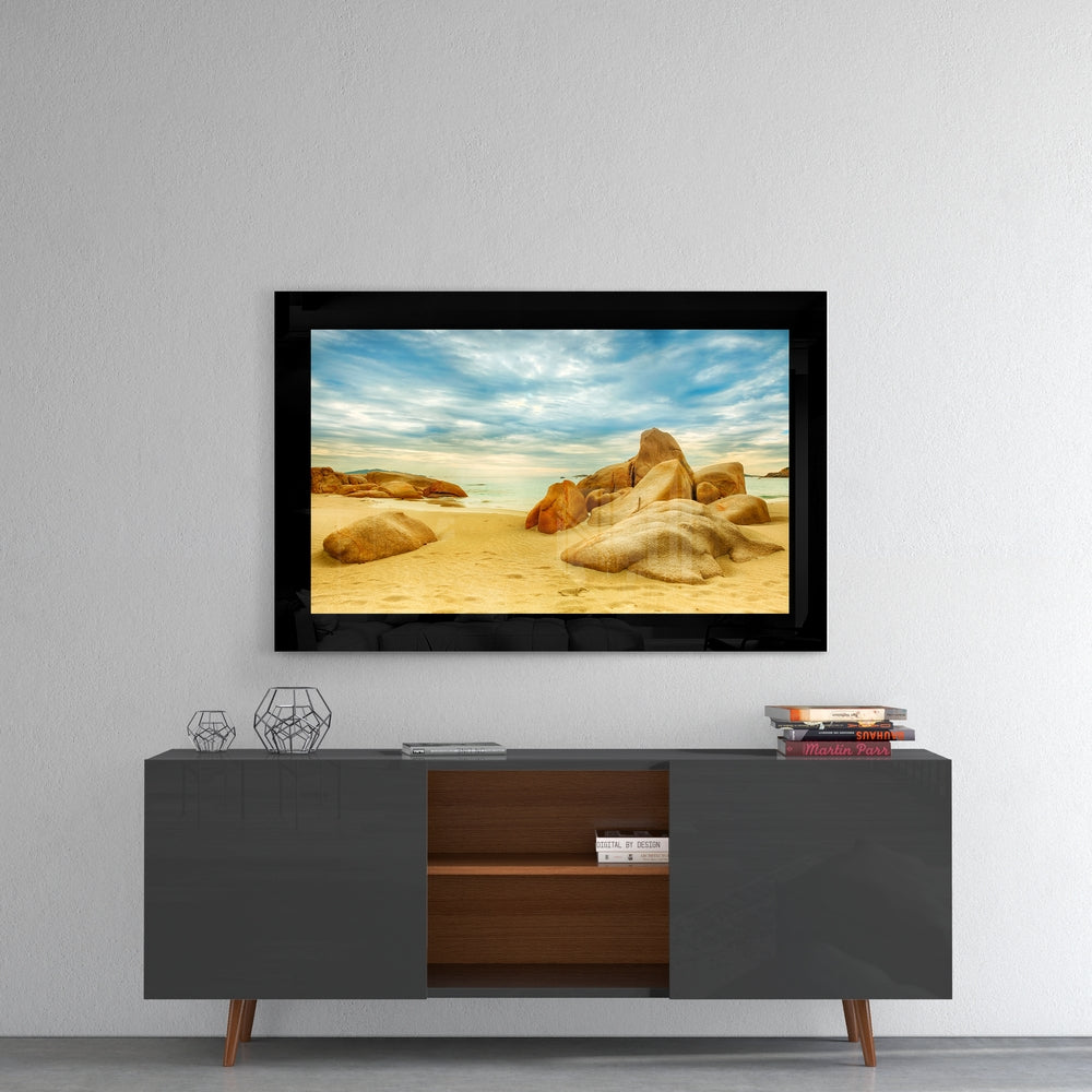 Black-framed glass wall art displaying a sandy beach with golden boulders and blue sky, mounted above a modern console table.