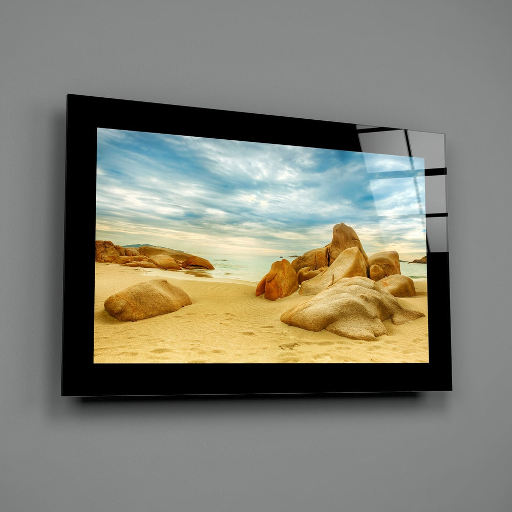 Black-framed glass wall art displaying a sandy beach with golden boulders, turquoise water, and cloudy sky, mounted on gray wall.