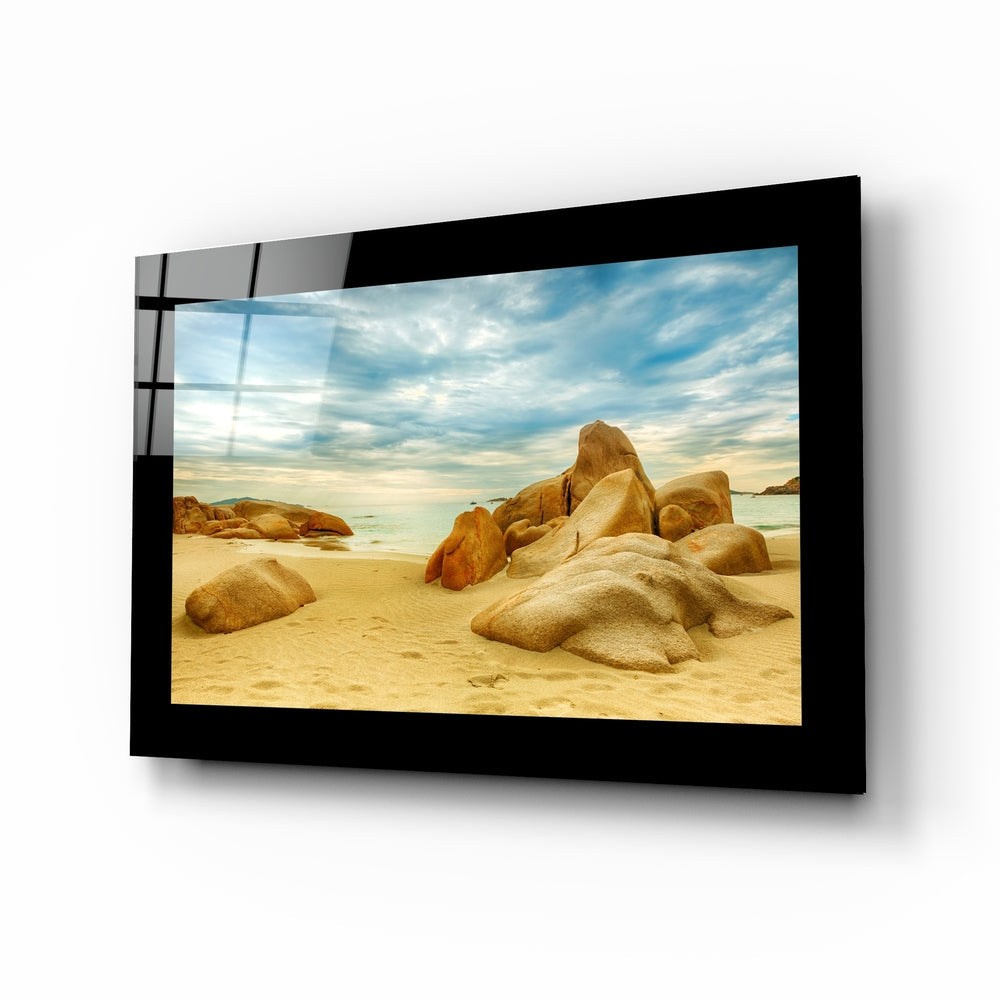 Black-framed glass wall art displaying a sandy beach with golden boulders and blue ocean horizon, mounted at an angle.