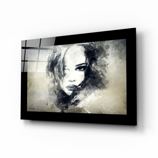 Black-framed glass wall art displaying a monochromatic watercolor portrait of a woman with ink splashes, angled perspective view.
