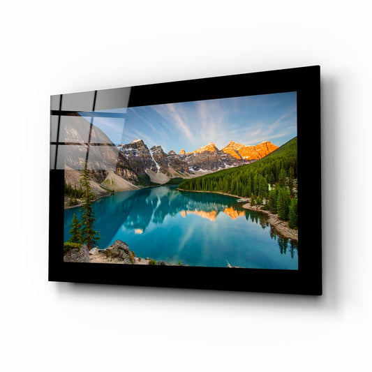 Black-framed glass wall art displaying a turquoise alpine lake surrounded by snow-capped mountains and evergreen forests, mounted at an angle.