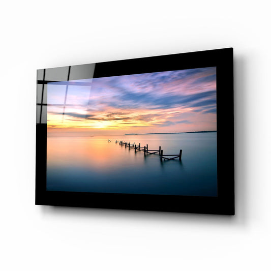 Black-framed glass wall art displaying a serene sunset over calm water with wooden pier posts, mounted at an angle against white background.
