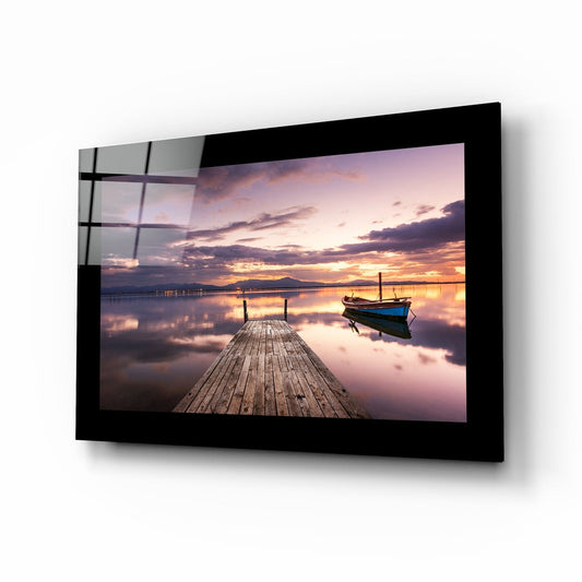 Glass wall art featuring a wooden dock extending toward a moored blue boat at sunset with purple and orange sky reflections.