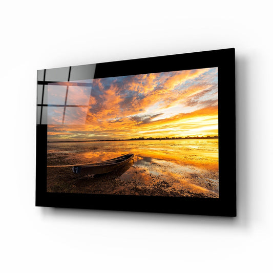 Black-framed glass wall art displaying a sunset landscape with boat on beach, mounted at an angle showing reflective surface and shadow.