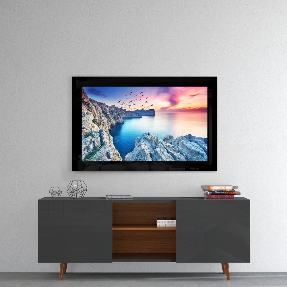 Glass wall art displaying a coastal sunset scene with rocky cliffs, blue water, and flying birds mounted above a mid-century modern TV console.